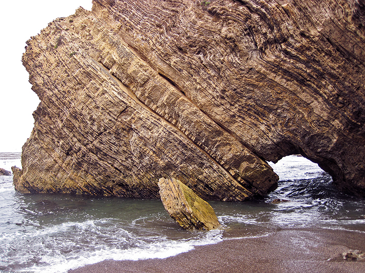 large, striated rock with small arch extrudes from the waterline